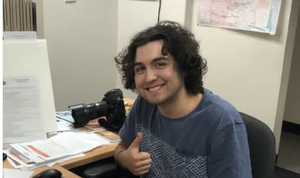 Young man with curly hair sits at an L-shaped desk. He wears a blue t-shirt. In the background are white walls, a map and a doorway. On the desk are papers, a camera, a computer keyboard and mouse.