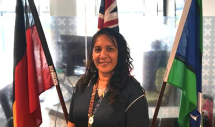 Indigenous woman with long black hair wearing black clothing stands in front of the Aboriginal, Australian and Torres Strait Islander flags. In the background is a glass wall behind which is an office.