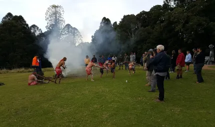 Indigenous dancers in full traditional dress perform next to a fire on a grassed area in front of onlookers. In the background are tall trees.