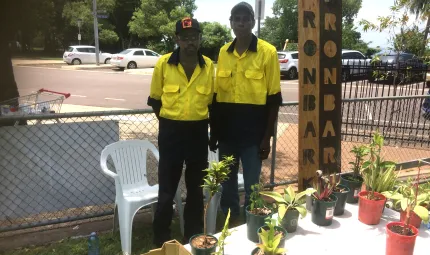 Two Aboriginal men standing outside in front of a table with pot plants and fruit. In the background there are trees and cars.