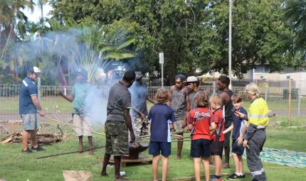 Group of young men stand around a fire placing long sticks over the fire from which smoke arises. In the background are trees, a fence and a light pole.