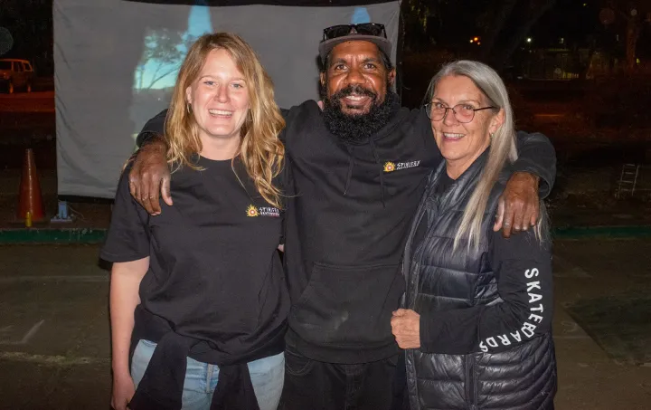 Two women and a man stand shoulder to shoulder. All are dressed in dark clothing. The background is dark except for a pale screen that displays a projected image.