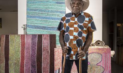 A man wearing a blue, pink, brown and white patterned shirt is standing in front of a collection of brightly coloured artwork. He is wearing a hat and is holding two, long carved sticks.