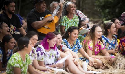 A group of people sit on chairs or on the ground and most wear colourful clothing. One man holds a guitar while some others hold papers. Most have their mouths open. In the background are trees.