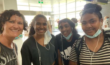 One adult woman and three young women stand shoulder to shoulder in a large room with windows and other people. Some have masks on their faces.
