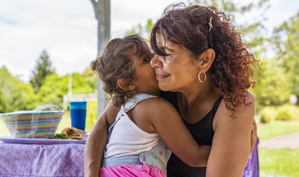 Woman in black top hugs a young girl in pink top. In the background is a table and trees.