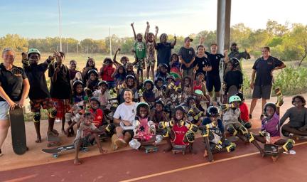 A group of young people and adults stand and sit on a hard surface. Some wear helmets and others hold skateboards. In the background is grass and trees and poles.