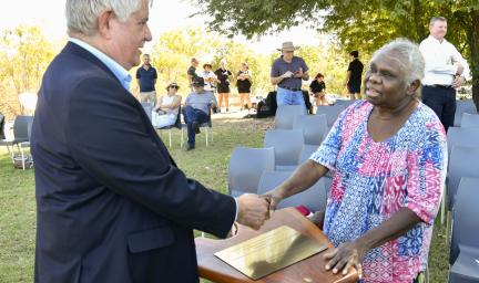 A man with grey hair and wearing a blue coat shakes hands with a woman with white hair and wearing a blue, pink and white dress. Under their hands is a podium and a gold engraved plate. In the background are chairs and people and trees.